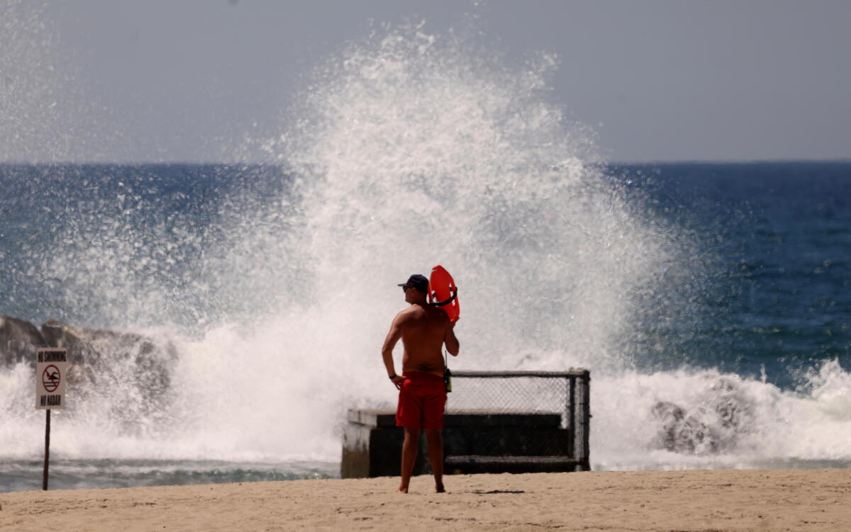 A lifeguard in red shorts holding a board in one hand stands on the beach watching a large wave crash onto the shore.